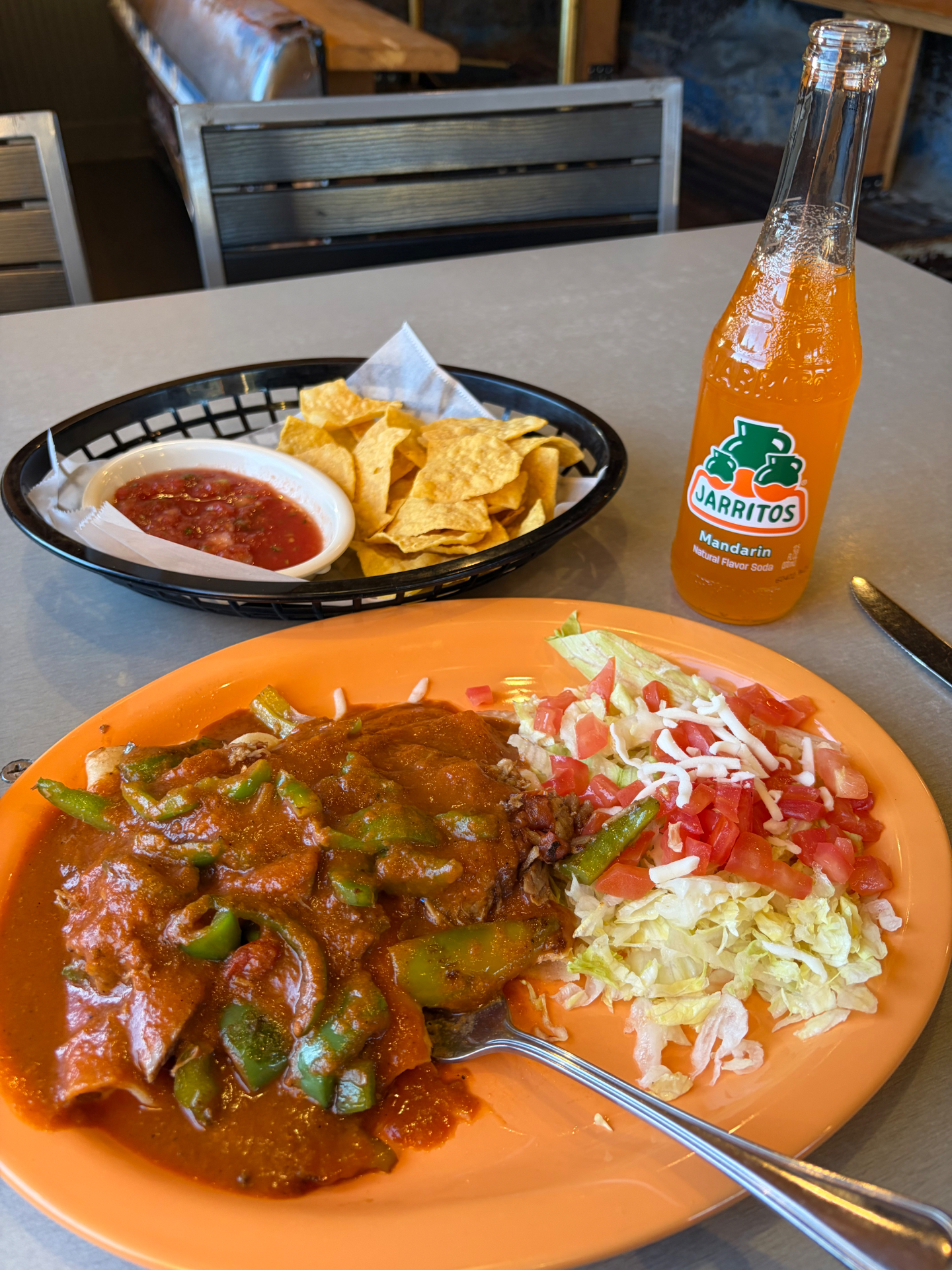 A plate of enchiladas smothered in red sauce with rice, lettuce and tomatoes, chips and salsa, and a bottle of Jarritos mandarin soda
