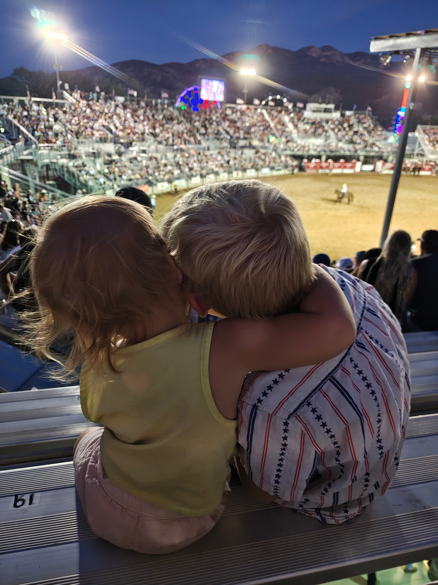 Two young children sitting close together on aluminum bleachers watching the Ute Stampede Rodeo in Nephi, Utah at dusk, with the arena lights and mountains behind them